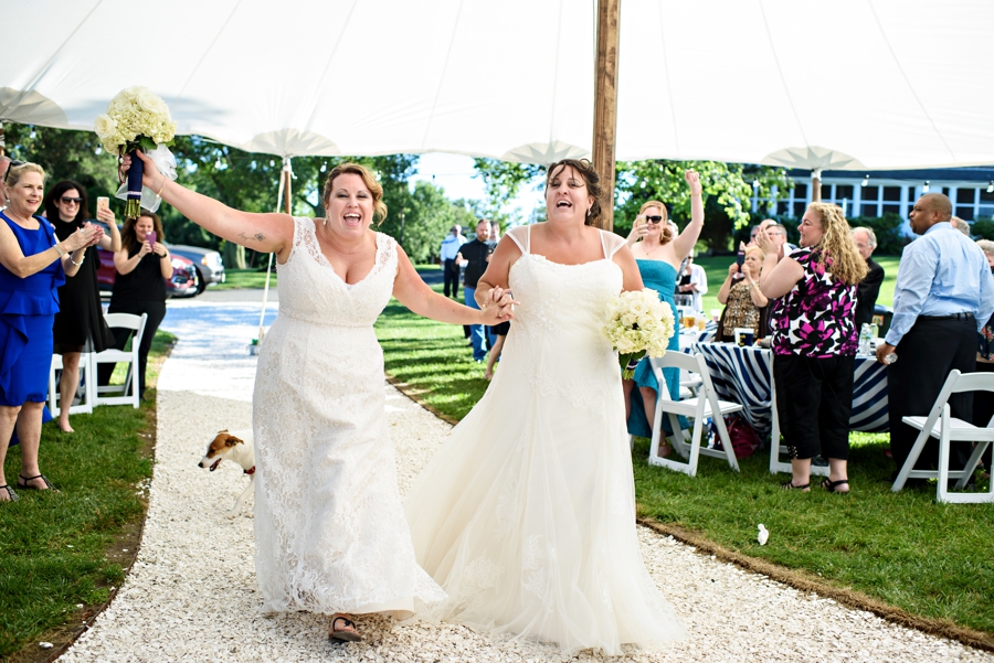 Two brides dance their way into their wedding reception under a giant white tent.